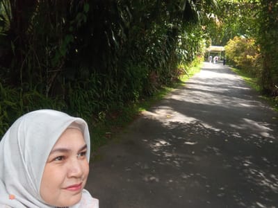 The image captures a woman wearing a white hijab, posing for a selfie in what appears to be a park or garden in Suka Makmur, Indonesia. She is positioned on the left side of the frame, looking to her right. Behind her, a paved pathway stretches into the distance, lined with lush green foliage on both sides. Several people are visible walking along the path in the background, and a light-colored structure, possibly a pavilion or canopy, is situated further down the path. The scene is illuminated by dappled sunlight filtering through the trees, casting shadows on the pathway.