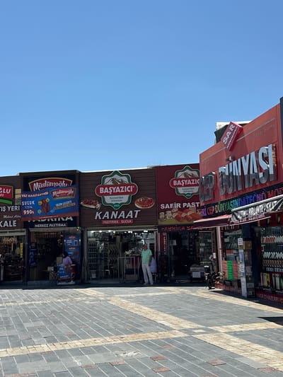 In the city of Kayseri, Turkey, a street scene unfolds under a clear blue sky. The foreground features a patterned pavement made of dark gray and light tan rectangular bricks. Beyond the pavement, a row of shops presents a vibrant facade of advertisements and signage.
One prominent shop displays a large banner for "Müslümoğlu" featuring images of cured meats and the text "YAZIN LEZZETİ MÜSLÜMOĞLU İLE BAŞLAR" (Summer's taste begins with Müslümoğlu). Adjacent to it, a red sign for "BAŞYAZICI" with an image of mountains reads "Kayseri'den Sofranıza 1953" (From Kayseri to your table 1953). Below this, a sign for "KANAAT" advertises "PASTIRMA - SUCUK" (cured meat - sausage).
Further to the right, a large, illuminated sign spells out "GÜNAYSI" in bold silver lettering, with smaller text above and below it. A colorful LED sign runs horizontally across the facade of this shop. Another shop on the far right displays a wide variety of items, including what appear to be phone accessories or electronics, arranged on shelves and in boxes.
Several individuals are visible. A person in a light green shirt and white pants stands near a shop entrance. Another person, dressed in a pink top and dark pants, is seen inside a shop, interacting with a display. A motorcycle is parked in front of one of the establishments. Inside the shops, various products, including strings of cured meats, are displayed. The overall impression is a bustling commercial street in Kayseri.