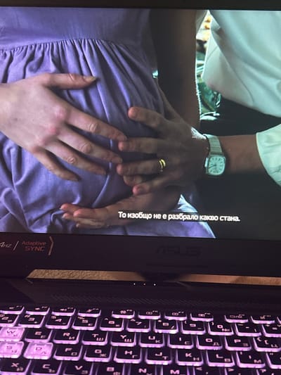 The image displays a close-up view of an open laptop in the city of Resilovo, Bulgaria, showing a scene on its screen and a portion of its backlit keyboard. The laptop is an ASUS model, indicated by the logo on the screen bezel, and features a purple-lit Cyrillic keyboard layout. The screen exhibits "4Hz Adaptive SYNC" technology and shows a still from a video, possibly a movie or television show.
The scene on the screen depicts the mid-section of a person with a visibly pregnant abdomen, clad in a light purple, gathered dress. Two sets of hands are positioned on the belly. One set of lighter-toned hands, presumably belonging to the pregnant individual, is spread across the abdomen. Over these, a second set of hands, appearing slightly darker, gently cups the belly. The individual associated with the second set of hands wears a plain gold wedding band on the left ring finger and a silver-toned square-faced watch with a metal link bracelet on the left wrist. A portion of a white collared shirt, belonging to this second individual, is visible on the right side of the screen. White Bulgarian subtitles are present at the bottom of the screen, reading: "То изобщо не е разбрало какво стана." (Translation: "It didn't understand what happened at all."). The background of the on-screen scene is out of focus, showing indistinct shapes and colors.