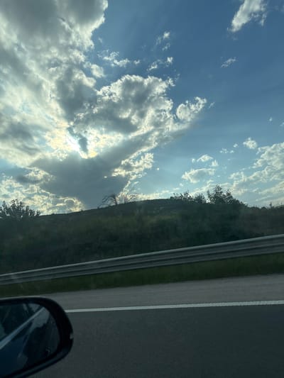 The image, taken from inside a moving vehicle, shows a view of the sky and a roadside in Kipra, Bulgaria. The sky is filled with white and gray cumulus clouds illuminated by sunlight. Directly to the right, a green hillside or embankment runs alongside a gray asphalt road. A metal guardrail is located between the hillside and the road. The right side of the image shows a portion of the car’s side mirror and the road surface with lane markings. No people are visible in the frame. The overall environment suggests daytime travel on a rural or semi-rural road.