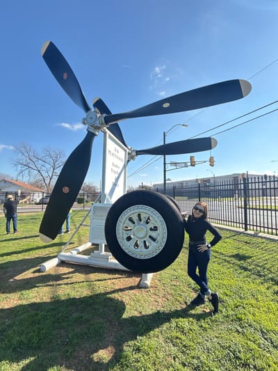 A woman in black clothing and sunglasses poses on a large aircraft tire mounted on a white stand. Behind her is a multi-blade propeller and a sign that reads "B-36 Peacemaker Bomber Propeller." The scene is outdoors, with a blue sky, green grass, and a chain-link fence in the background. Street lights and buildings are visible in the distance. The image appears to be rotated.