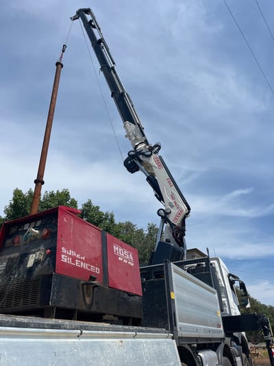 An exterior medium shot shows a large red and black industrial generator on the bed of a truck, positioned in the city of Castiglione delle Stiviere, Italy. A white crane is actively lifting a rusty metal pipe from above the generator. The pipe is suspended by a cable attached to a metal hook. The generator has "SUPER SILENCED" printed on its side in white letters and "MOSA GE 22 VSX" in red. The truck also has a silver cargo bed. The sky is blue with scattered white clouds, and green trees are visible in the background.