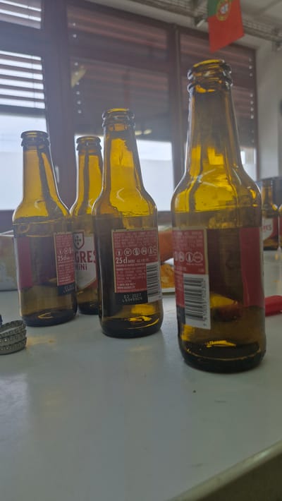 In Uetendorf, Switzerland, four amber glass beer bottles are arranged on a white, reflective surface. Three bottles are in the foreground, and one is slightly behind and to the right. The bottles are mostly empty, with some residual liquid visible at the bottom. Each bottle has a label with text and nutritional information. The bottle on the far left has a red label with white text. The second bottle from the left has a white label with a crest and text. The third bottle from the left has a label with text that includes "25cl BIÈRE ALC. 4.8% VOL." and a barcode. The bottle on the far right also has a red label with white text and a barcode. A Portuguese flag is visible in the background, hanging from a cord. The background also shows a window with horizontal blinds and a partially obscured outdoor scene. Several metal bottle caps are stacked to the left of the bottles.