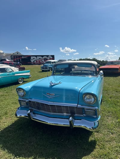 A fleet of classic cars are parked on a grassy field under a bright blue sky with scattered clouds, possibly at an outdoor event or car show. In the foreground, a light blue two-door convertible with a white top is prominently displayed, facing the viewer. Its chrome bumper and grille gleam in the sunlight. To its left, another vintage car, teal in color, with a white top and chrome accents, is visible. Further in the background, a light blue vintage van or SUV and a bright orange car are parked. Behind the vehicles, a large black trailer with white lettering and graphics is visible, as are some trees and buildings, one of which is white with pillars. The environment suggests a daytime outdoor gathering, potentially in a rural or suburban setting within the city of Fairburn, United States. No individuals are clearly discernible in the immediate foreground or interacting with the vehicles.