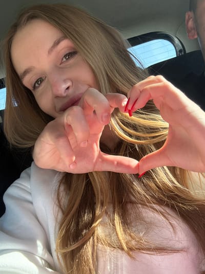 A female subject with light-colored, long hair and light brown eyes is depicted in a close-up, selfie-style image. She smiles faintly towards the camera. Her hands are positioned in the foreground, forming a heart shape; her right hand displays long, pointed nails with red polish, while her left hand has natural, unpolished nails. She wears a light-colored garment, possibly a white hoodie. In the upper right background, a partial view of a male's ear is visible. Behind the subjects, a car's rear window with defroster lines reveals a clear blue sky, indicating an outdoor, daylight setting within a vehicle. The scene is located in the city of Praga, Montenegro.