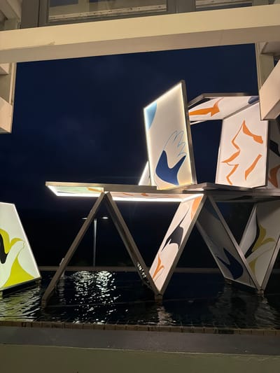 The image shows an illuminated abstract sculpture composed of geometric shapes and panels with colorful graphic designs. The sculpture is situated outdoors, over a body of dark water that reflects the lights and the artwork. The background is a dark night sky, with some faint lights visible in the distance, possibly streetlights. The sculpture itself features white panels with stylized graphic elements in blue, orange, yellow, and black. These panels are arranged in an unstable, house-of-cards-like structure, some of which appear to be lit from within. The structure is supported by metallic, triangular frames. In the foreground, the edge of a tiled surface is visible, leading to the water. The overall impression is modern and artistic, likely a public art installation in an urban setting.