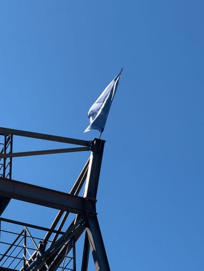 A low-angle, full shot captures a blue flag fluttering against a clear, bright blue sky. The flag is attached to the top of a dark, angular metal structure that resembles part of a construction or industrial framework. This structure, with its stark beams and bolted joints, occupies the lower left and center of the frame, silhouetted against the sky. The scene appears to be outdoors, with the strong blue of the sky suggesting a sunny day. The metalwork is dark and industrial, indicating a construction site or a permanent industrial installation. There are no people or animals visible in the image. The overall impression is one of a bright, open sky contrasted with the geometric, man-made structure. This scene is of the new metalwork frame on the Yelets embankment, designed to hold the flag of Russia.