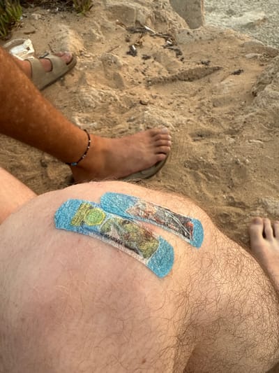 A high-angle shot reveals a person seated outdoors on sand, displaying hairy legs. One leg wears a sandal and a beaded anklet. On the other leg, two adhesive bandages, possibly featuring Marvel characters, cover the knee area. The ground consists of a mix of sand and pebbles, with some vegetation. The overall setting suggests a casual, outdoor environment.
