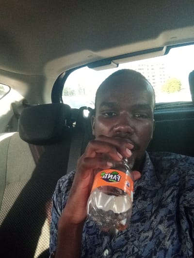 An adult male, with dark skin and short dark hair, is depicted from the chest up, seated in the back of a vehicle. He wears a blue, patterned, short-sleeved collared shirt with visible buttons. His right hand holds a clear plastic bottle, labeled "FANTA" with an orange graphic, to his mouth, suggesting he is drinking from it. The bottle contains a clear liquid. To his left, a dark-colored fabric seat with a headrest and a visible seatbelt is present. Through the vehicle's rear window, a brightly lit outdoor scene is visible, featuring a road or open area on the left and a multi-story building with green foliage on the right. The scene occurs during daylight hours within the city of Makadara/Mji Wa Kale ward, Kenya.