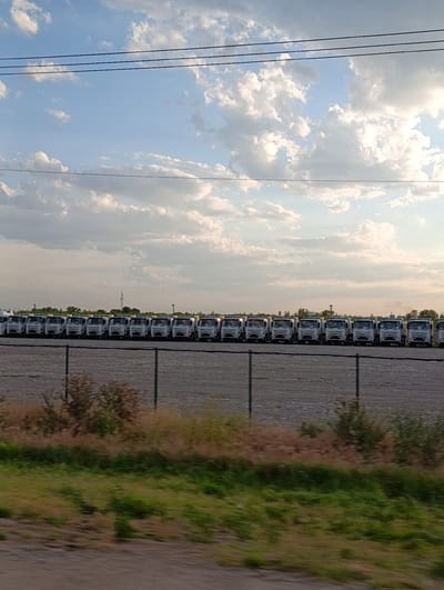 An eye-level, medium shot captures a row of identical white trucks parked in a lot under a cloudy sky. The trucks are aligned in a single file, creating a repetitive pattern. In the foreground, a chain-link fence runs horizontally across the image, separating the lot from a strip of dry grass and some bushes. Beyond the fence, the trucks are parked on a gravel surface. In the distance, trees and a transmission tower are visible against the horizon. The overall scene suggests an industrial or storage area.