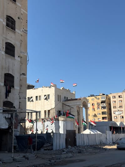 In Jabalia, Palestinian Territories, a building damaged by conflict stands on the left, its concrete facade marked with numerous bullet holes and debris. On the right, several buildings are visible, including a yellow multi-story structure and a lighter-colored building with arched windows. The scene is further characterized by Palestinian flags displayed prominently on poles atop some buildings and in front of a row of white tents. The ground is littered with rubble and debris, suggesting recent destruction. A person in a red shirt walks away from the camera in the lower left quadrant of the image, near a dark tarp and some makeshift structures. A silver car is parked on the right side of the frame. The sky is clear and blue.