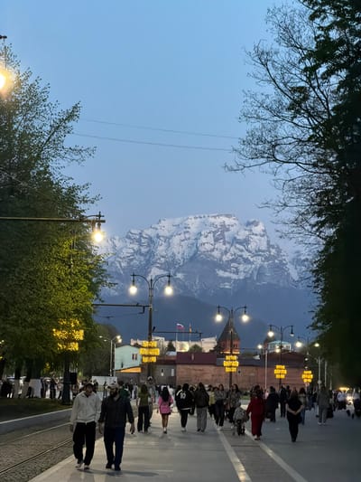 The image presents a street scene in Chermen, Russia, at dusk. A snow-capped mountain forms a backdrop in the distance. A row of illuminated lampposts lines the street. People are walking on the sidewalk along the tramway track. Trees flank the street. Some branches are bare while others are covered in leaves. The sky is light blue. A Russian flag is visible near the buildings.