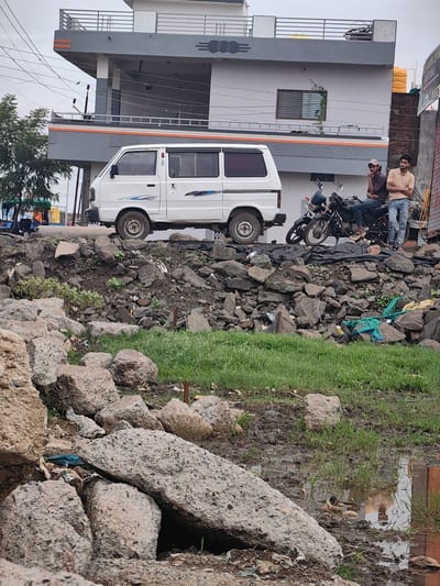 The image depicts a street scene in Padli, India, with a focus on a white van parked on a raised platform. In the background, a multi-story building with a modern facade stands out. Two men are visible near a motorcycle, with one man sitting on the motorcycle and the other standing beside it. The foreground is characterized by a rocky terrain with scattered green grass and some muddy patches, suggesting a somewhat unkempt environment. The overall impression is of an urban fringe or developing area.