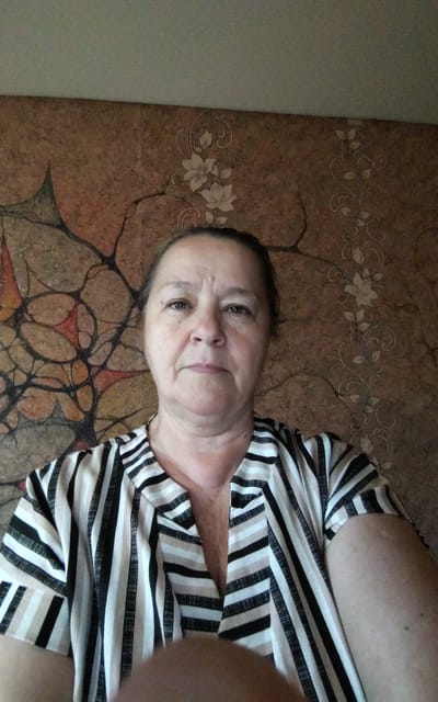 A medium-shot, eye-level selfie captures a middle-aged woman with fair skin and brown hair, pulled back. She is looking directly at the camera with a neutral expression. She is wearing a striped V-neck shirt with black, white, and beige stripes. The background is a textured wall with a floral wallpaper pattern. The image appears to be taken indoors, possibly in a residential setting in Zheleznogorsk-Ilimsky, Russia.