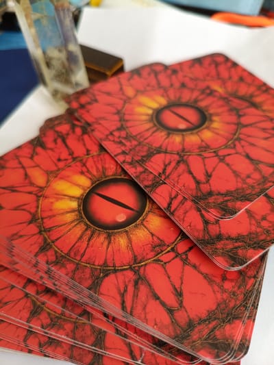This is a close-up, high-angle, static shot of a deck of red cards with a stylized eye design, possibly a tarot deck, fanned out and overlapping on a white surface. The eye design is predominantly red with yellow and black accents, creating a fiery, reptilian appearance. In the background, out of focus, are various objects including a clear crystal, a dark rectangular object, and a blue surface. There are no people or animals present. The scene is indoors and appears to be a still life arrangement. There is no information available to place this image in Nizhnekamenka, Russia.