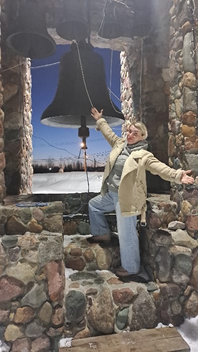 An individual stands within a stone bell tower in Avdot'ino, Russia, during a winter evening. The structure is constructed from diverse, irregularly shaped stones, some partially covered with snow. Three large, dark metal bells are suspended from the top of the tower's opening, with a prominent central bell and two smaller bells on either side. The individual, appearing female with blonde hair, is positioned between the stone columns, facing the camera. She wears a light beige shearling-lined jacket, a grey hooded sweatshirt, light blue jeans, and brown shoes. Her arms are outstretched and slightly raised, with hands open, and a faint smile is visible. Her right hand is near a white rope connected to the largest bell. She stands with her left foot on a lower stone ledge and her right foot elevated on a higher stone step. A small light bulb hangs below the largest bell, and string lights are visible along the right stone column. The ground within and around the bell tower is covered in a layer of snow. Beyond the tower, under a dark blue sky, a snow-covered landscape with sparse trees and distant structures or utility poles is visible. A blue rope is coiled on a stone ledge to the left of the individual. A wooden plank is situated on the snowy ground at the bottom of the frame.