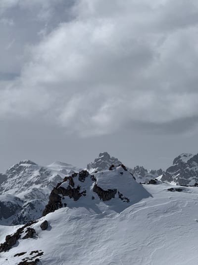 The scene displays a vast, snow-covered alpine landscape in Puy-Saint-André, France. The foreground consists of an expansive snowfield marked by subtle wind-sculpted textures and scattered dark rock outcrops; faint ski tracks are discernible in the lower-left quadrant. A prominent rocky peak, partially covered in snow and casting a shadow, dominates the midground. Beyond this, successive ranges of rugged, snow-capped mountains recede into the distant haze. The sky is uniformly overcast with dense layers of grey clouds, providing diffuse illumination across the entire scene. No human figures or artificial structures are visible.