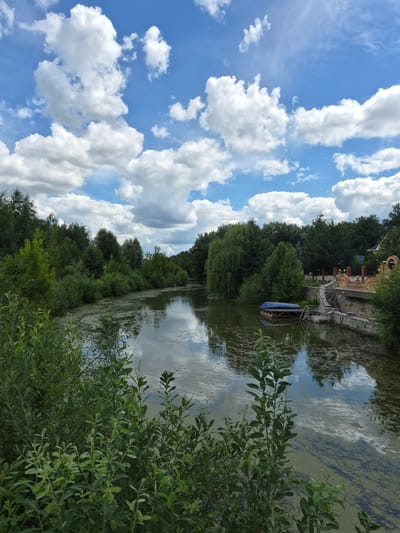 A scenic view of a tranquil river winding through a lush green forest. The sky above is a brilliant blue, dotted with fluffy white clouds that cast gentle reflections on the water's surface. The riverbanks are densely lined with trees, their branches reaching out towards the water. A small, blue-covered boat is docked on the right side of the river, near a stone-built structure with steps leading down to the water. The water itself is calm, with patches of green algae floating on top, creating an almost painterly effect. The overall scene evokes a sense of peace and natural beauty, possibly from the outskirts of Vlast Truda, Russia, given the presence of a more developed structure on the bank, suggesting proximity to a settlement or resort.