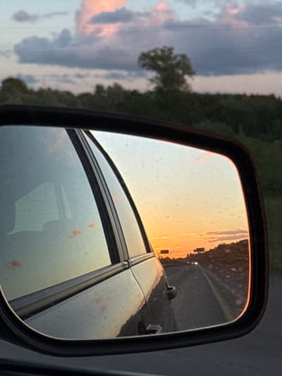 The image is a close-up of a car's side-view mirror reflecting a sunset over a highway. The mirror shows a road stretching into the distance, with cars on it and signs on the side. The sky in the reflection is filled with warm orange and yellow hues from the setting sun, with some clouds tinged pink. The foreground of the image shows the car's side and the frame of the mirror. The car appears to be dark-colored. Outside the mirror, the sky is a dusky blue with scattered clouds, and a silhouette of trees and a single tree can be seen on the horizon. The environment appears to be rural or suburban. There are no people visible in the image. The scene depicts a journey during sunset, with the reflection in the mirror offering a view of the road ahead and the sunset. The location is not identifiable as Nagornaya, Russia, or any specific city.
