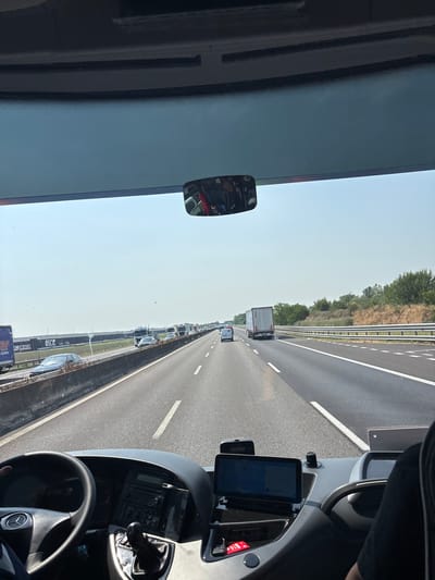 The image shows a view from inside a vehicle, likely a bus or truck, on a highway. Outside, there is a traffic jam of vehicles, including a white box truck. The road has multiple lanes and a barrier on the left side. There is a green area with trees in the distance. Inside the vehicle, there is a steering wheel with a Mercedes-Benz logo, a dashboard with a navigation device, and a rearview mirror showing a reflection. The windshield is tinted. While it's impossible to confirm the precise location as Parma, Italy without further data, the highway infrastructure and general vegetation are consistent with the region.