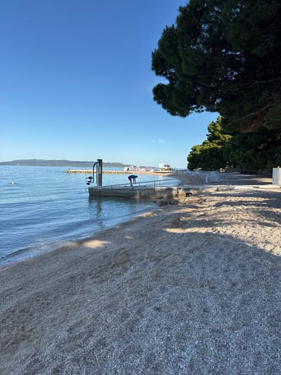 The image depicts a sunny day at a beach in Makarska, Croatia. The foreground is dominated by a pebble beach with gentle waves lapping at the shore. In the mid-ground, a concrete pier extends into the clear blue water, where a person in a blue shirt and red shorts is performing a stretching exercise. Further out in the sea, a rocky jetty is visible with a few people on it, and in the distance, inflatable water attractions are set up. The background features a treeline of dark green pine trees casting shadows on the beach, and a hazy mountain range under a clear blue sky. Several white lounge chairs are stacked on the beach, suggesting a recreational area. The scene is peaceful and bathed in natural light.