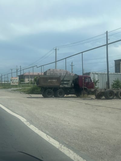 The image captures a roadside scene in Ayntap, Armenia, during daytime with a partly cloudy sky. The photograph is oriented vertically, rotated 90 degrees counter-clockwise.

In the foreground, a paved road or asphalt surface with a white lane marking is visible. Dominating the middle ground are two trucks parked alongside the road. The truck closer to the viewer is a dump truck with a load of what appears to be bricks or rectangular construction materials. On its side, the text "094 27 8863" is clearly legible. Behind this truck, a red truck, possibly an older model, is visible.

Multiple utility poles with electrical wires stretch across the scene, indicating an established infrastructure. In the background, beyond the trucks and utility lines, there are several buildings, some appearing residential and others possibly industrial, along with some green foliage. A billboard or large sign is partially visible in the distance, with the word "ARMENIA" prominently displayed. The overall setting suggests a semi-urban or industrial area. No specific activity or people are discernible in the image.