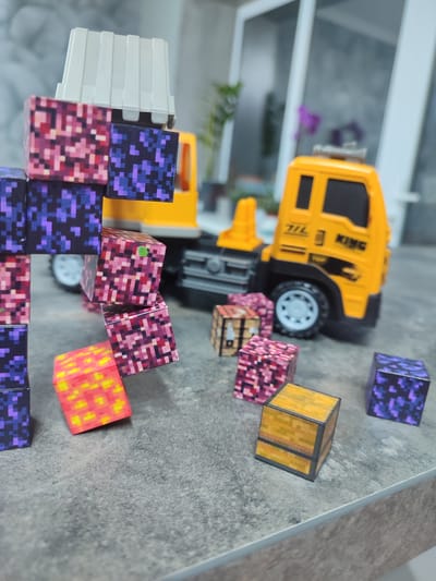 The image shows a close-up, eye-level view of a toy truck and several small pixelated blocks. The truck is yellow with black accents and a white cargo container. The blocks are of various colors and patterns, with some appearing to be stacked. The scene is set on a grey, textured surface, possibly a countertop. In the background, a blurry window with vertical blinds and some greenery is visible. There is no indication of the city of Grigoriopol, Moldova, or any specific location within it.