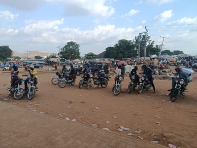 The image depicts an outdoor scene at a marketplace in Gbessa, Nigeria. Several motorcycles are parked on a dirt lot littered with debris and plastic waste. The marketplace is crowded with people and stalls covered with umbrellas, indicating potential commercial activity. A mix of vehicles including cars and motorcycles, are visible within the market area. Overhead power lines are visible. The landscape includes trees, distant hills, and a sky with clouds. The foreground shows a paved area adjacent to the dirt lot. Individuals are seen standing or sitting on the motorcycles.