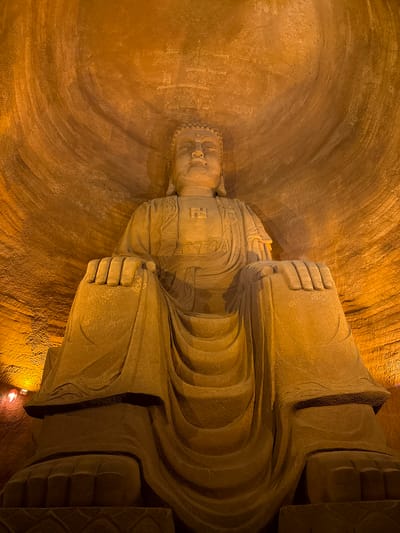 This image depicts a large, stone-carved statue of a reclining Buddha, situated within a cave or grotto. The statue dominates the frame, occupying most of the space. The Buddha's head is positioned towards the left, and the body extends to the right. The statue features a solemn facial expression, and the garments appear to be intricately carved. A swastika is visible in front of the chest. The surrounding cave walls have a rough texture, possibly indicating the marks of chiseling or carving. The scene appears to be dimly lit, with warm tones dominating the color palette. A light source is visible near the bottom right, providing some illumination to the lower portion of the statue. This statue is located in Shizhong, China.