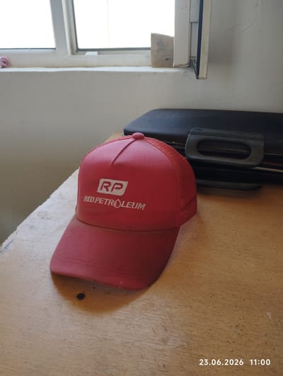 An indoor, eye-level, medium shot shows a red baseball cap with a white logo that reads "RP RED PETROLEUM" resting on a light brown, wooden table. Behind the cap, a black, hard-shell case with a silver handle is visible. The background consists of a white wall and a window with white trim. The lighting suggests it is daytime. The date and time stamp "23.06.2026 11:00" is present in the bottom right corner. This image was taken in Sokuluk, Kyrgyzstan.