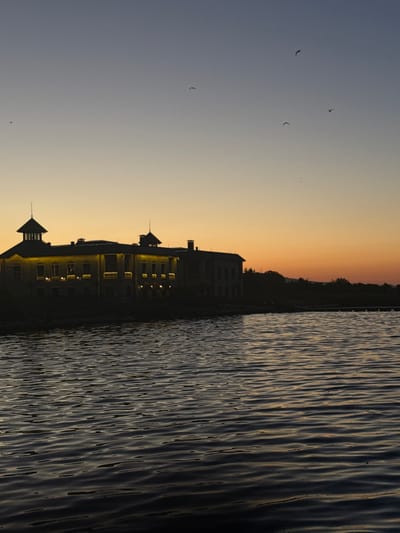 The image depicts a twilight scene over a body of water, with a silhouetted building on the horizon. The sky transitions from a pale blue at the top to a warm orange near the horizon, suggesting the sun has recently set or is about to rise. Several birds are visible as small dark shapes against the sky.
The building is illuminated by warm yellow lights from within, highlighting its architectural features. It appears to be a substantial structure with at least two prominent towers. The water in the foreground is rippled, reflecting the dim light of the sky. The overall impression is one of tranquility.
The prompt states this scene is located in the city of Казашко, Bulgaria. Given the visual elements, this could be a waterfront building, such as a hotel, restaurant, or a historical edifice, near the coastline or a large river. The presence of the water and the style of the building could align with coastal or riverside developments in Bulgarian towns.