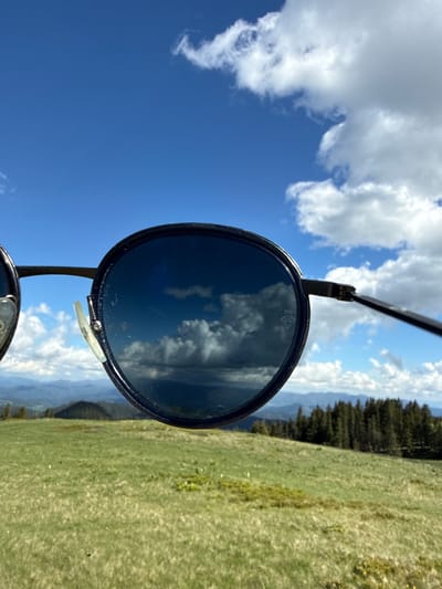 An outdoor medium shot shows sunglasses held up to the camera, obscuring the upper portion of the frame. The sunglasses have round, dark lenses and thin black frames. Reflected in the left lens are white, puffy clouds against a bright blue sky. The right lens also reflects clouds, some appearing darker and more dramatic. A small diamond shape with the letters "PH" is visible etched onto the lower right lens.

The background, visible through and around the sunglasses, depicts a hilly landscape under a partly cloudy sky. A grassy, green meadow occupies the foreground and midground, dotted with small yellow flowers. A line of dark green pine trees forms a dense forest along the slope of a distant hill. Beyond the trees, layers of hazy blue mountains recede into the distance. The overall impression is of a clear, sunny day in a mountainous or high-altitude area. There is no indication of the city of Gela, Bulgaria in this image.