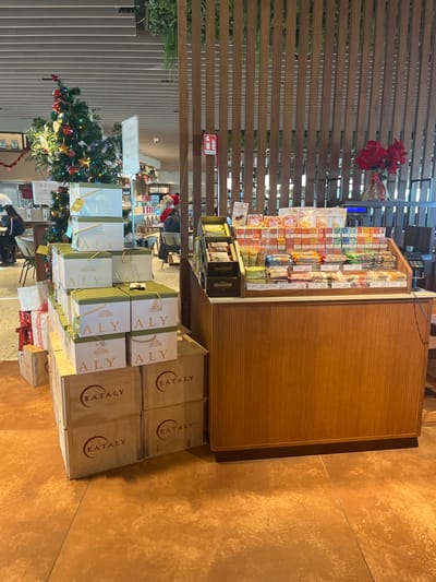 This indoor shot, oriented vertically, captures a bustling commercial space, likely a specialty food market or store, decorated for the holiday season. In the mid-ground, a long, light wooden display counter stretches across the frame, laden with numerous packaged food items, including various candies, chocolates, and what appear to be gourmet snacks, meticulously arranged in rows. Below and to the right of this counter, several light wooden crates are stacked, prominently featuring the red "EATALY" logo on their sides, suggesting an Italian food retail outlet.
To the left of these crates, a collection of elegant gift bags, primarily in white with gold and green accents, are stacked high. The letters "ALY" are visible on some of these bags, possibly part of a larger brand name. Beneath these bags, red and white gift-wrapped boxes are also visible, contributing to the festive atmosphere.
In the background, a fully decorated Christmas tree, adorned with colorful ornaments and lights, stands prominently to the left, further emphasizing the holiday setting. Behind the tree and display counter, a slatted wooden partition or wall creates a textured backdrop. A red sign, likely indicating a fire extinguisher, is mounted on this partition. A few people can be faintly seen in the deeper background, with one person wearing a red Santa hat. The floor is covered in large, textured reddish-brown tiles. The overall scene suggests a vibrant retail environment, possibly in Dorno, Italy, given the Eataly branding and the context provided.