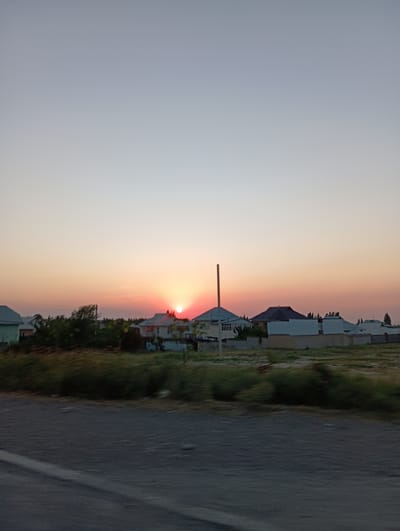 The image shows a sunset scene in the village of Sadirbay, Kyrgyzstan. The sky is a gradient of pale blue to warm orange and pink hues as the sun dips below the horizon. In the mid-ground, several residential buildings with varying roof designs are visible, interspersed with greenery and trees. A tall, slender pole stands to the left of the frame, near one of the houses. The foreground consists of a blurred asphalt road, suggesting the photo was taken from a moving vehicle. The overall atmosphere is calm and serene, indicative of the end of a day in a rural setting.