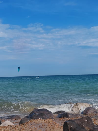 A kite surfer is seen on the ocean, with a bright blue kite against a backdrop of a cloudy blue sky. The sea is a vibrant turquoise, with gentle waves lapping at the shore. The beach in the foreground is covered in pebbles and dark, wet rocks. In the distance, on the horizon, a line of wind turbines is visible. The scene suggests a breezy, sunny day, likely during daylight hours, with the activity of kite surfing indicating a recreational or sporting event. The location, Worthing, United Kingdom, provides context for the coastal setting. No text is visible in the image.
