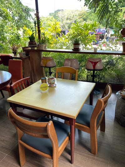 This image depicts the interior of a restaurant in Bang Sare, Thailand, with a view of lush greenery through a large window. In the foreground, a light-colored square table is set with two wooden chairs and another chair partially visible. On the table, there's a small decorative pot, a metal cup, and a yellow sign with Thai text. In the background, through the window, a variety of plants are visible, along with some outdoor structures and a distant hill. The seating area features several unique bar stools shaped like buttocks, one with red underwear. The overall atmosphere suggests a quirky and relaxed dining environment.