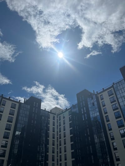 A low-angle, outdoor, vertical shot captures a modern apartment building under a bright, partly cloudy blue sky with the sun shining brightly. The building's facade is a contrast of light-colored panels and dark, reflective glass windows arranged in a grid pattern. Some sections of the building are darker, appearing to be more heavily glazed. The top of the building features black railings along balconies. Wispy white clouds are scattered across the sky, with the sun's rays creating a lens flare. The lower portion of the image is dominated by the building, angled upwards towards the sky. The scene is devoid of any people or discernible human activity, presenting a clean and architectural view of urban development in Prigorodny, Russia.