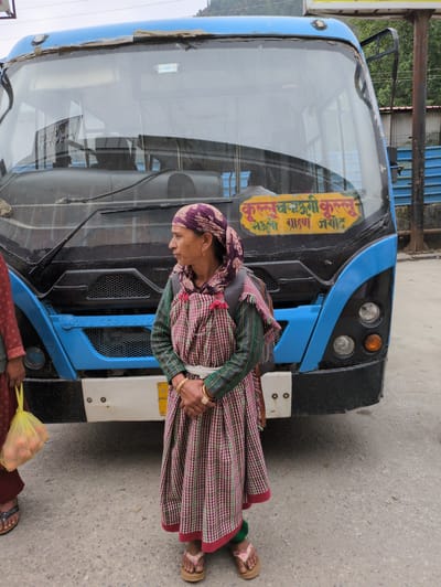 A woman in a patterned headscarf and a long, checkered dress stands in front of a blue and black bus. She has her hands clasped in front of her and wears a backpack. She is facing to her right, looking away from the camera. Another person's legs are visible on the left side of the image, holding a yellow bag filled with fruit. A yellow sign with writing in Hindi is affixed to the front of the bus. The scene appears to be in Kullu, India, based on the writing on the bus. The ground is paved with concrete, and a metal fence can be seen in the background.