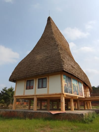 The image shows a traditional Vietnamese house on stilts, located in Đà Giang, Vietnam. The house features a steep, conical roof thatched with brown material, and white walls adorned with panels depicting cultural scenes or figures. The house is elevated on light-colored wooden pillars, creating an open space underneath. A wooden walkway connects to the side of the house. The house is surrounded by green grass, with trees visible in the background under a sky with scattered clouds.