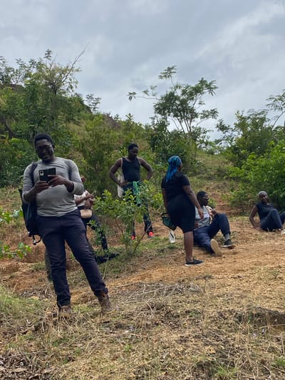 A group of five people are outside in a rural location with trees and dry grass. One man in the foreground is standing, wearing a grey shirt, black pants, brown shoes, glasses, and a backpack. He is holding a black smartphone and looking at the camera. Further back, another person is standing with their hands on their hips, wearing a black tank top and floral-patterned shorts. One woman is standing, wearing a black top, black skirt, and a blue headscarf. Two other people are sitting or lying on the ground. The location is likely Ganawuri, Nigeria, based on the prompt. The sky is overcast.