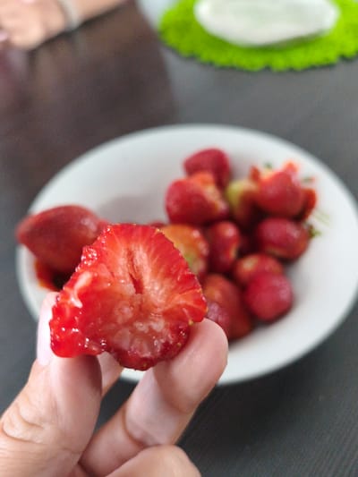 A close-up, eye-level shot shows a hand holding a sliced strawberry in the foreground, with a white bowl of strawberries in the background. The sliced strawberry is halved, revealing its internal structure, which appears somewhat soft and juicy with visible seeds. The hand holding it is lightly colored and appears to be composed of fingers and a thumb. The bowl behind it contains several whole strawberries, some of which are brighter red than the sliced one. The overall scene is set against a dark, textured surface, likely a table, with a hint of a green, circular object in the upper right corner and a blurred background suggesting an indoor environment. There is no indication of the city of Brodi, Latvia, in this image.