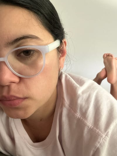 The image displays a female individual situated in the city of Mar del Plata, Argentina. She is positioned indoors, likely lying prone or reclining. The left side of her face is prominent, featuring a neutral expression, dark hair, and light-colored, rounded spectacle frames. Her left eye, with a brown iris, is visible through the lens. A small, silver-toned hoop earring is present in her left earlobe. She is wearing a light pink, short-sleeved t-shirt with visible stitching on the shoulder. Her bare feet, soles visible and flexed upwards, are present in the upper right portion of the frame. The background consists of a plain, light-colored surface.