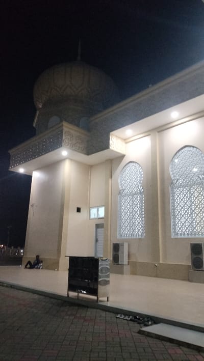 This nighttime photograph captures a well-lit mosque or religious building in Mee Tanjong Usi, Indonesia. A large, patterned dome is visible against the dark sky on the left. The building's light-colored facade features a balcony-like structure with intricate geometric designs, illuminated by recessed lights. Two prominent arched windows on the right side display decorative latticework, and below them are two outdoor air conditioning units. A smaller, rectangular window is visible on the left side of the building.
In the mid-ground, a dark, multi-tiered shoe rack is positioned on a paved area, with a small pile of shoes on the ground nearby, suggesting a common entrance practice for mosques. To the left, a lone individual is seated on the paved ground next to the building wall, appearing to be resting or waiting. The ground in the foreground is paved with interlocking bricks, transitioning to a smoother, lighter-colored surface closer to the building. The artificial lighting creates a warm glow on the building's exterior, contrasting with the dark night. The scene suggests a tranquil atmosphere, possibly before or after evening prayers or a community gathering. No discernible text is visible.