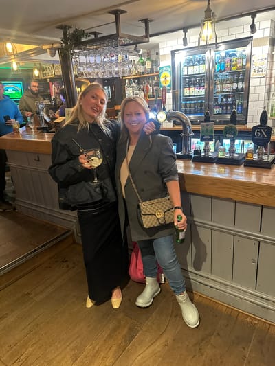 The image captures two women posing in what appears to be a pub. The woman in the foreground, to the left, is holding a martini glass with a lime wedge. She wears a dark denim jacket over what looks like a black dress or skirt. Her blonde hair is shoulder-length. The woman behind her is holding a green bottle and has a light-brown Gucci-style handbag slung over her shoulder. She wears a light-grey coat, jeans, and white rubber boots.
The scene is a traditional pub setting with a wooden floor, bar, and shelving displaying a wide selection of alcohol. Behind the women, there's a display refrigerator filled with various bottled drinks. Beer taps and vintage-style lighting fixtures are also visible.
The pub is likely located in St Albans, United Kingdom, based on the brand labels visible on the beer taps and the general aesthetic of the pub, which is consistent with British pubs.