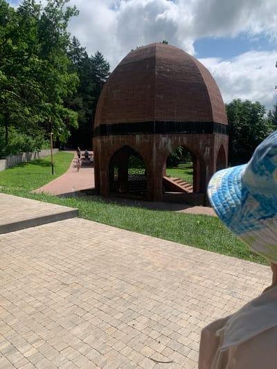 In Arbuzov-Baran, Russia, a circular brick structure with arched openings stands prominently in a park on a cloudy day. The structure has a domed roof and a dark band encircling its midsection, adorned with decorative metalwork. Inside, a set of stairs leads upwards. The surrounding area features a paved walkway, lush green grass, and mature trees. Several people, including a child in a pink dress, are visible walking along the path, some approaching the structure. The foreground is dominated by a beige brick pathway, and the back of a person wearing a blue and white patterned hat is in the lower right corner of the frame.