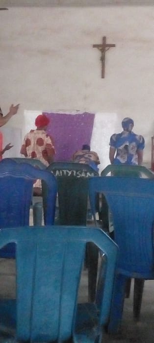The image, taken indoors, depicts a gathering of people, likely in a religious or community setting. The scene is dominated by a plain, light-colored wall on the left, upon which a wooden crucifix, depicting Jesus, is mounted.

In the foreground and mid-ground, several individuals are seated on blue and green plastic chairs. The chairs are arranged in rows, suggesting an organized assembly. Most of the people visible are seen from behind or from the side.
One person, seated on a green plastic chair, wears a blue and white patterned top and a blue head covering.
Another person, seated on a similar green chair, wears a light-colored top with dark polka dots and a red head covering.
Further to the right, a row of blue plastic chairs is visible, some of which appear to be occupied, though the occupants are largely obscured.
In the bottom left corner, an arm and a hand are visible, raised as if in participation or gesture.
A patch of purple fabric or decoration is visible on the wall behind some of the seated individuals.

On the back of one of the green chairs, partial text is visible, appearing to spell out "AITYTSAN". The overall lighting is consistent with an indoor environment, possibly illuminated by natural light from unseen windows or by artificial lighting. The atmosphere appears to be one of quiet gathering or worship.