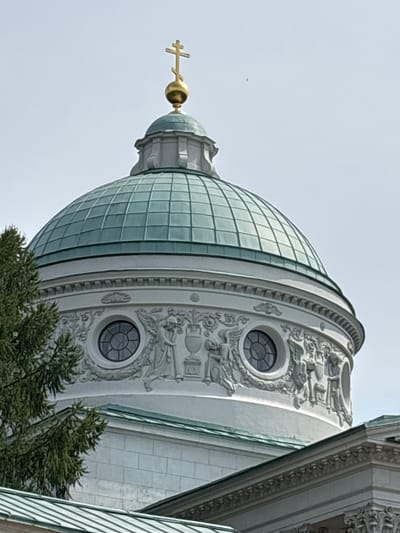 This is a close-up, low-angle shot of the dome of the Church of the Archangel Michael in the village of D.N.P. Arkhangelskoye, Russia. The dome is teal-colored and segmented, topped by a golden Orthodox cross on a golden orb. The white stone base of the dome features intricate relief sculptures, including circular windows and figures resembling angels or cherubs, and classical urns. A large green tree branch obscures the left side of the image. The sky is overcast and pale blue. Below the dome, architectural elements of the church, including a white stone wall with a tiled roof, are visible.