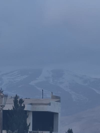 The image displays an outdoor scene in Qudsaiya, Syria, featuring a large expanse of sky above a modern building and some vegetation. The sky, which dominates the upper and left parts of the frame, is a soft blue-grey with distinctive, wispy white cloud patterns stretching across it, indicating a partly cloudy or overcast day.

In the bottom right quadrant, a multi-story building with a contemporary design is visible. It is constructed from light-colored material, possibly concrete or stone, featuring sharp, geometric lines and rectangular cut-outs or windows that appear dark due to shadow or dark glass. A thin, dark pole or antenna extends vertically from an upper section of the building. Dark green foliage, likely from trees, is visible in the foreground, partially obscuring the lower levels of the structure.

The overall lighting suggests daytime, with a soft, diffused light, indicating no direct harsh sunlight. There is no discernible human activity or visible text in the scene. The image provides a static view of the architectural and natural elements within this specific location.
