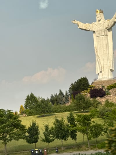 A statue of Jesus Christ, wearing a golden crown, stands tall against a pale sky with scattered clouds. The statue is white, with arms outstretched. It is situated on a hill, with a stone retaining wall and lush greenery, including trees and bushes, surrounding its base. In the foreground, a grassy slope descends, lined with various trees. At the bottom of the image, several trash cans are visible along a pathway. The scene is located in Swiebodzin, Poland, home to the Christ the King statue.