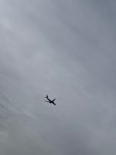 Against a backdrop of mottled gray skies, an airplane ascends. Positioned in the lower right quadrant, the aircraft is seen from below, its fuselage and wings silhouetted. Engine exhaust is visible behind the wings. The sky is predominantly overcast with varying shades of gray and hints of cloud texture. The airplane occupies a relatively small portion of the frame, emphasizing the vastness of the sky.